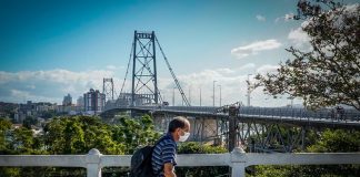 homem usando máscara passa em calçada com parapeito; ao fundo ponte hercílio luz - regras estaduais contra Covid em SC