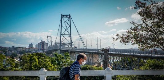 homem usando máscara passa em calçada com parapeito; ao fundo ponte hercílio luz - regras estaduais contra Covid em SC