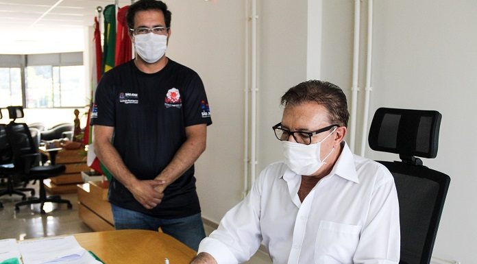 Sentado em uma cadeira preta, o prefeito Orvino usa camisa branca de manga comprida, além de óculos de armação preta e máscara branca. Ele assina um convênio que destina mais de R$ 1 milhão para a área do esporte em São José. Próximo a ele, em pé, um dos representante da área acompanha a assinatura, ele usa óculos de grau, máscara branca e camiseta preta.
