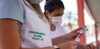 Funcionária usa relógio e jaleco branco, com escrita em verde que diz secretaria municipal de saúde Palhoça-SC. Ela está com um frasco e uma seringa de vacina. A vacinação dos maiores de 70 anos inicia nesta semana em Palhoça.