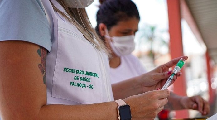 Funcionária usa relógio e jaleco branco, com escrita em verde que diz secretaria municipal de saúde Palhoça-SC. Ela está com um frasco e uma seringa de vacina. A vacinação dos maiores de 70 anos inicia nesta semana em Palhoça.