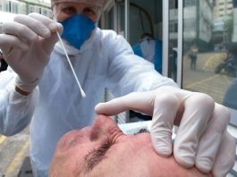 Procon notifica planos de saúde para autorização imediata de testes de covid close no rosto de idoso com cabeça inclinada; profissional de saúde todo trajado de epi aproxima com a mão grande cotonete ao nariz, para fazer teste; mão do profissional sobre a cabeça do idoso