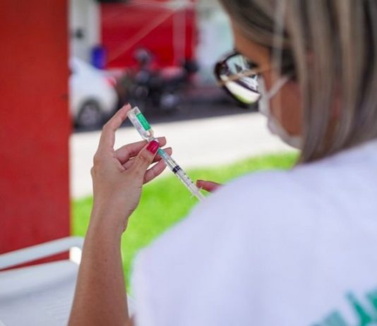 Vacinação: SC tem 409 mil imunizados e recebe na sexta novo lote com 129 mil doses Profissional veste roupa branca, óculos e máscara. Ela está de costas manipulando uma seringa e um frasco de vacina. Nesta semana a vacinação em Palhoça chega aos idosos com mais de 79 anos.