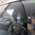 profissional de saúde aplica vacina no braço de senhora no banco do carona; reflexo do vidro do carro na frente