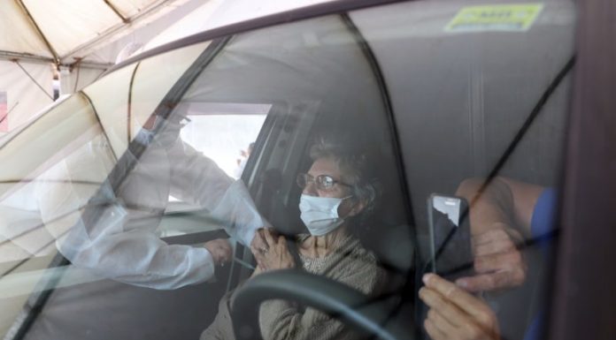 profissional de saúde aplica vacina no braço de senhora no banco do carona; reflexo do vidro do carro na frente