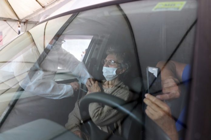 profissional de saúde aplica vacina no braço de senhora no banco do carona; reflexo do vidro do carro na frente