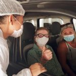 idosa faz sinal de positivo para a foto dentro de carro com outra mulher ao lado e profissional de saúde na janela - São José e Florianópolis começam a vacinar idosos de 78 anos; em Palhoça, 77 anos