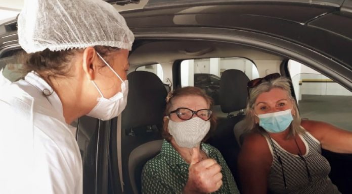 idosa faz sinal de positivo para a foto dentro de carro com outra mulher ao lado e profissional de saúde na janela - São José e Florianópolis começam a vacinar idosos de 78 anos; em Palhoça, 77 anos