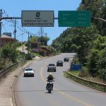 motoclista passando sob placa da sc 283, indicação de distância das cidades de seara, itá e chapecó e outros carros na rodovia de pista simples