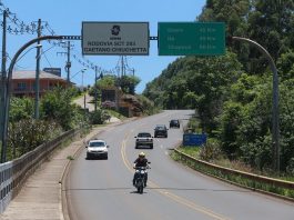 motoclista passando sob placa da sc 283, indicação de distância das cidades de seara, itá e chapecó e outros carros na rodovia de pista simples