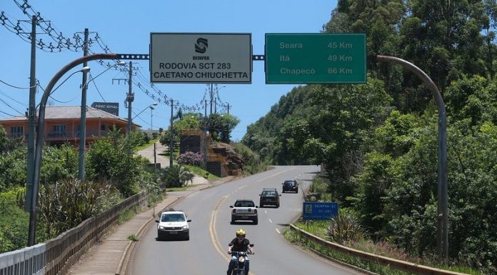 motoclista passando sob placa da sc 283, indicação de distância das cidades de seara, itá e chapecó e outros carros na rodovia de pista simples
