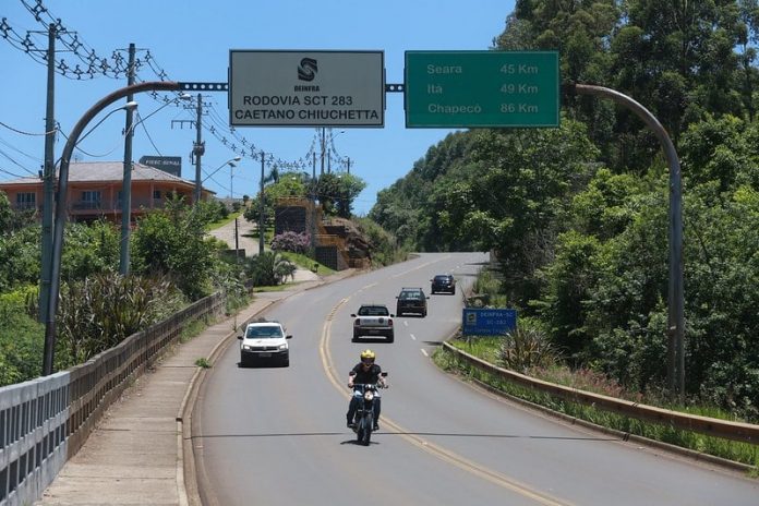 motoclista passando sob placa da sc 283, indicação de distância das cidades de seara, itá e chapecó e outros carros na rodovia de pista simples