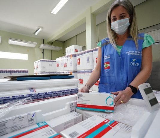 SC tem 2,1% da população vacinada, previsão de poucas doses em abril e mudança no lote funcionária usando máscara e colete confere caixas de doses de vacinas dentro de caixas de isopor