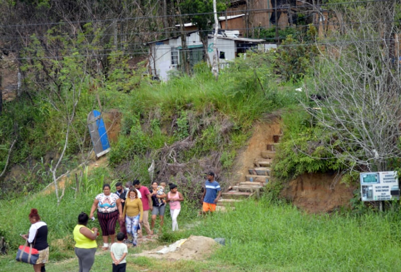 mulheres andando em várzea no pé de encosta com mata onde há algumas casas precárias de madeira