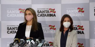 daniela reinehr e carmen zanotto usando máscara, em pé, em frente a bancada com microfones; ao fundo painel com logos do governo de santa catarina