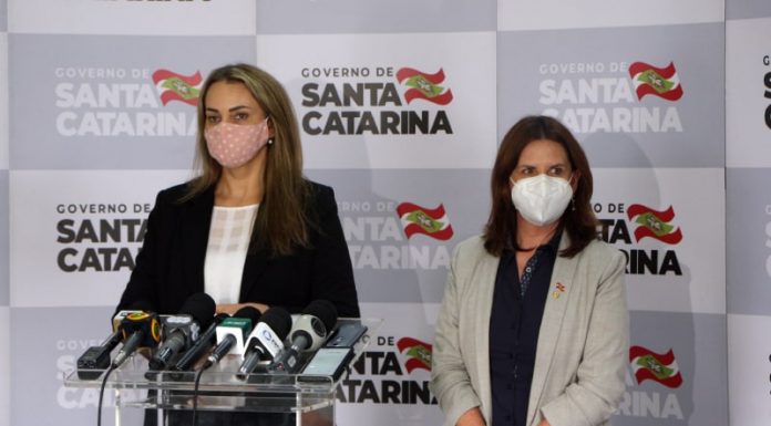 daniela reinehr e carmen zanotto usando máscara, em pé, em frente a bancada com microfones; ao fundo painel com logos do governo de santa catarina