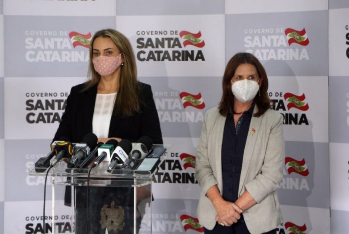 daniela reinehr e carmen zanotto usando máscara, em pé, em frente a bancada com microfones; ao fundo painel com logos do governo de santa catarina
