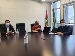 Em uma mesa grande, três pessoas sentadas, separadas por uma cadeira entre cada um, todos usam máscaras. Essa foi a reunião que chegou ao plano de vacinação nos profissionais da educação de SC.