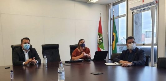 Em uma mesa grande, três pessoas sentadas, separadas por uma cadeira entre cada um, todos usam máscaras. Essa foi a reunião que chegou ao plano de vacinação nos profissionais da educação de SC.