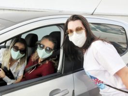 três mulheres usando máscara e óculos escuros posam para foto; duas estão dentro do carro e uma próxima, do lado de fora, em drive-thru de vacinação