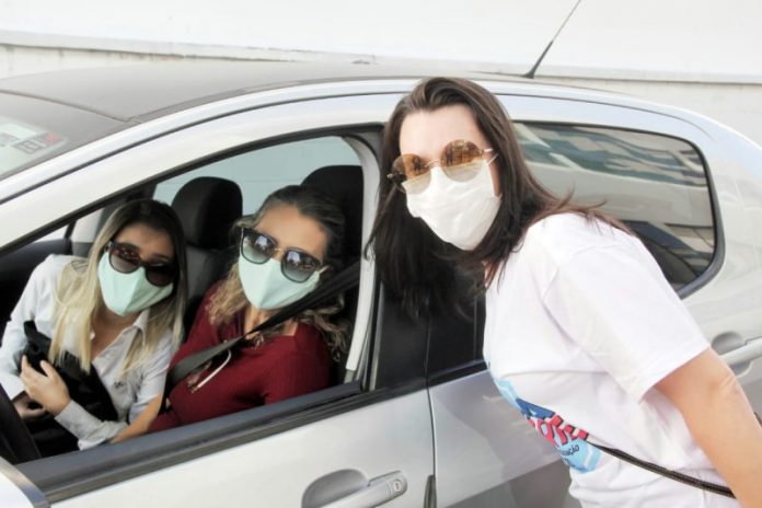 três mulheres usando máscara e óculos escuros posam para foto; duas estão dentro do carro e uma próxima, do lado de fora, em drive-thru de vacinação