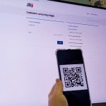 tela de um computador em página branca com logo de são josé na parte de cima, é o site de cadastro para vacinas do município, a frente da tela uma mão segura um celular com um qr code aberto.