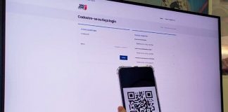 tela de um computador em página branca com logo de são josé na parte de cima, é o site de cadastro para vacinas do município, a frente da tela uma mão segura um celular com um qr code aberto.