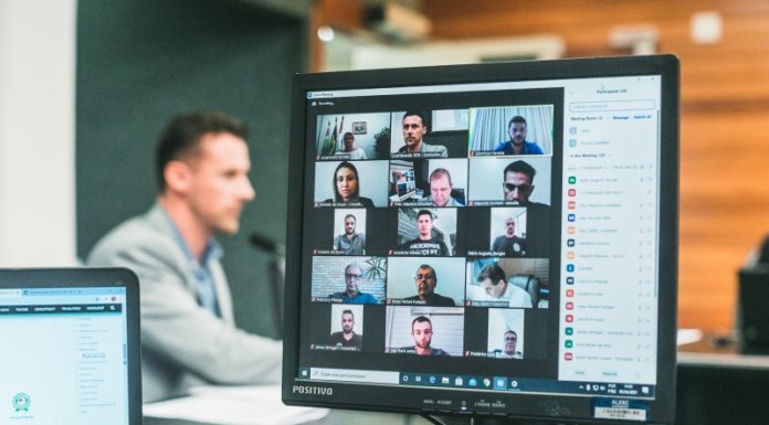 O foco da foto é em uma tela de computador que mostra 15 quadrados com o vídeo de participantes da audiência pública sobre o funcionamento das arenas de atividades esportivas de SC. Ao fundo o deputado que solicitou o encontro.