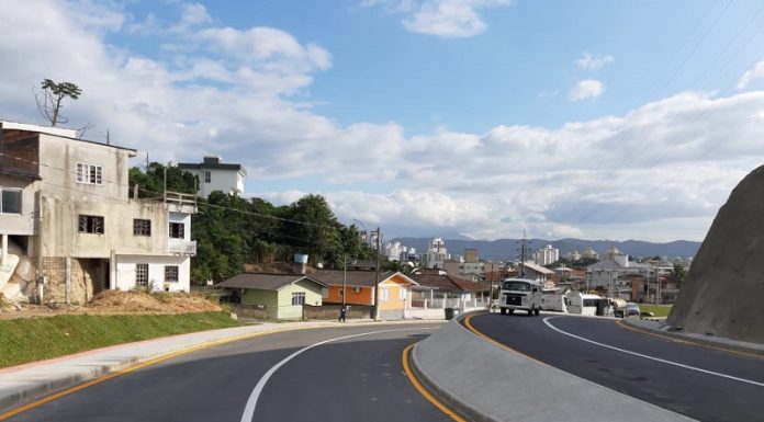 A foto mostras a avenida das torres em Palhoça, com asfalta novo e pista dupla. O céu é azul e ao fundo é possível ver algumas residencias.