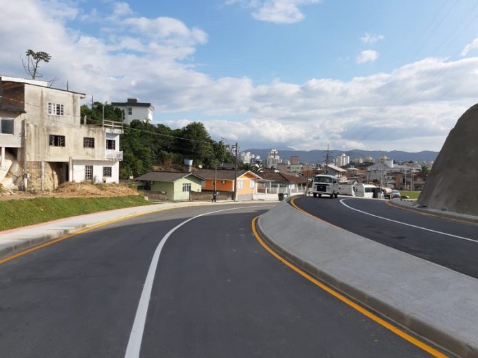 A foto mostras a avenida das torres em Palhoça, com asfalta novo e pista dupla. O céu é azul e ao fundo é possível ver algumas residencias.
