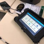 O equipamento Cellebrite, utilizado pelo IGP de SC, tem bordas pretas e em sua tela é possível ver quadrados brancos. A ferramenta está conectado por um cabo curso a um celular que aparece ao fundo da tela.