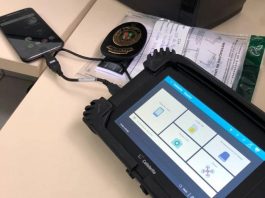 O equipamento Cellebrite, utilizado pelo IGP de SC, tem bordas pretas e em sua tela é possível ver quadrados brancos. A ferramenta está conectado por um cabo curso a um celular que aparece ao fundo da tela.