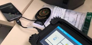 O equipamento Cellebrite, utilizado pelo IGP de SC, tem bordas pretas e em sua tela é possível ver quadrados brancos. A ferramenta está conectado por um cabo curso a um celular que aparece ao fundo da tela.