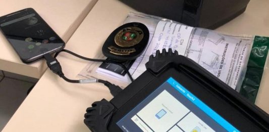 IGP inicia entrega de novos equipamentos israelenses Cellebrite O equipamento Cellebrite, utilizado pelo IGP de SC, tem bordas pretas e em sua tela é possível ver quadrados brancos. A ferramenta está conectado por um cabo curso a um celular que aparece ao fundo da tela.