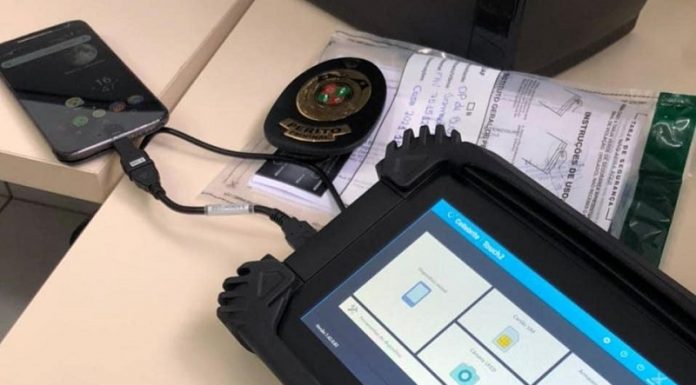 O equipamento Cellebrite, utilizado pelo IGP de SC, tem bordas pretas e em sua tela é possível ver quadrados brancos. A ferramenta está conectado por um cabo curso a um celular que aparece ao fundo da tela.