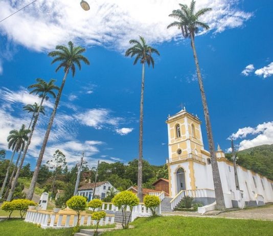 A foto com ângulo de baixo para cima mostra o céu azul e ensolarado com nuvens brancas, há árvores altas e é possível ver construções históricas. A prefeitura de palhoça organiza um concurso fotográfico em seu aniversário de emancipação.