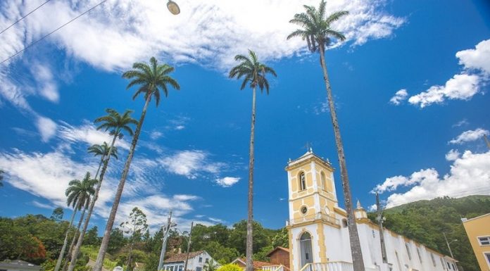 A foto com ângulo de baixo para cima mostra o céu azul e ensolarado com nuvens brancas, há árvores altas e é possível ver construções históricas. A prefeitura de palhoça organiza um concurso fotográfico em seu aniversário de emancipação.