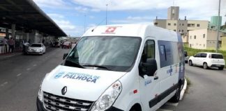 Ambulância branca com identificação em azul da prefeitura de Palhoça. O veículo está parado em frente a estação Palhoça, onde ocorre a vacinação do município.