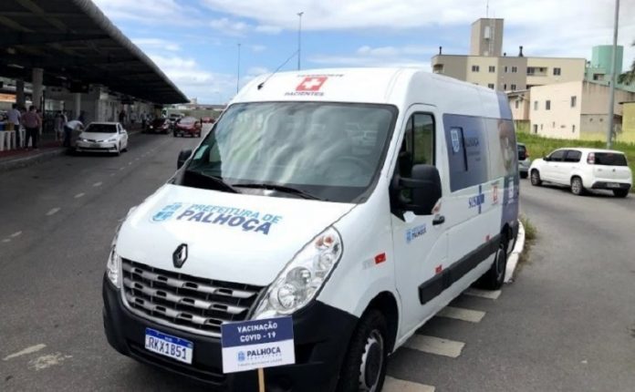 Ambulância branca com identificação em azul da prefeitura de Palhoça. O veículo está parado em frente a estação Palhoça, onde ocorre a vacinação do município.