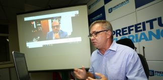 Gean Loureiro veste uma camiseta azul clara e óculos, ao fundo se vê inscrições que identificam a prefeitura de Florianópolis. Loureiro, que é presidente do consórcio nacional de vacinas, fala para a tela do computador e ao fundo se vê o diretor do instituto butantan, eles discutem o cronograma da coronavac.