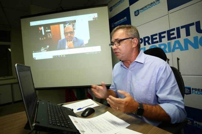 Gean Loureiro veste uma camiseta azul clara e óculos, ao fundo se vê inscrições que identificam a prefeitura de Florianópolis. Loureiro, que é presidente do consórcio nacional de vacinas, fala para a tela do computador e ao fundo se vê o diretor do instituto butantan, eles discutem o cronograma da coronavac.