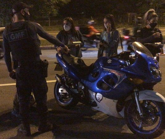 Guarda de Florianópolis faz blitz contra motos barulhentas A foto em período noturno mostra uma moto parada com dois guardas municipais e duas pessoas conversando com eles, todo usam máscaras. Ao fundo uma movimentação de moto e carros. A GMF passa a utilizar um aparelho para identificas as motos em alta velocidade e emissão de ruídos