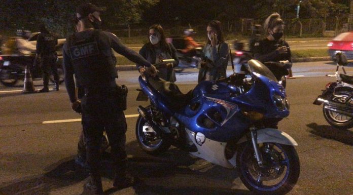 A foto em período noturno mostra uma moto parada com dois guardas municipais e duas pessoas conversando com eles, todo usam máscaras. Ao fundo uma movimentação de moto e carros. A GMF passa a utilizar um aparelho para identificas as motos em alta velocidade e emissão de ruídos