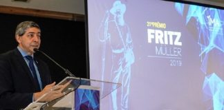 Um homem de terno e cabelo grisalho fala em frente a um microfone. Ao fundo, um telão mostra a capa do 21º prêmio Fritz Muller, do IMA.