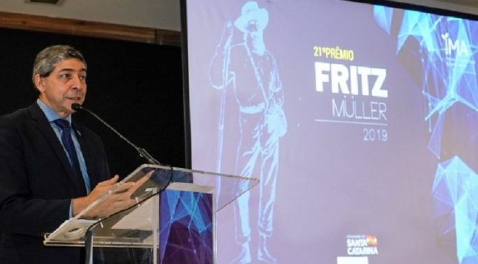 Um homem de terno e cabelo grisalho fala em frente a um microfone. Ao fundo, um telão mostra a capa do 21º prêmio Fritz Muller, do IMA.