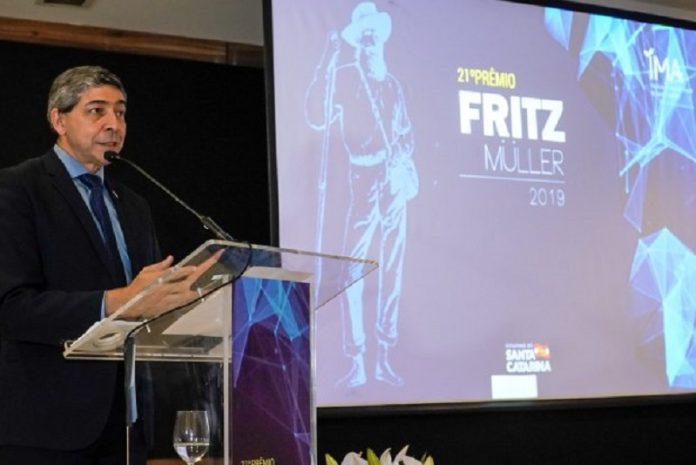 Um homem de terno e cabelo grisalho fala em frente a um microfone. Ao fundo, um telão mostra a capa do 21º prêmio Fritz Muller, do IMA.