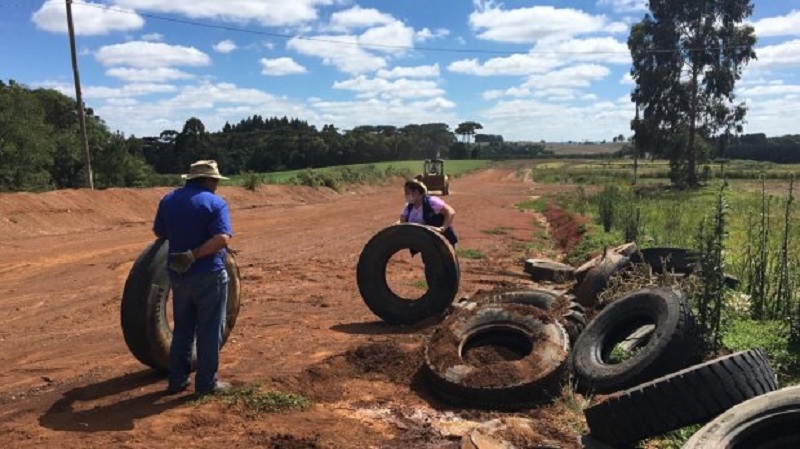 Em uma estrada de terra, duas pessoas mexem em pneus que parecem abandonados. O programa do IMA visa o gerenciamento correto de resíduos, como os pneus.