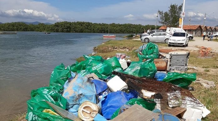 Na margem do Rio Cubatão sacolas de lixo empilhadas, ao fundo alguns carros.