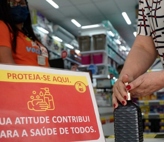 placa ao lado de álcool em gel em entrada de loja escrito sua atitude contribui para a saúde de todos - pessoa pegando álcool gel, funcionária de máscara ao lado, prateleiras ao fundo