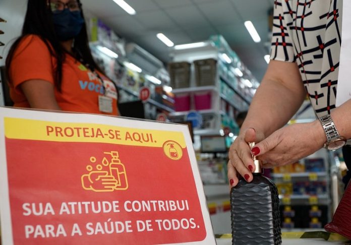 placa ao lado de álcool em gel em entrada de loja escrito sua atitude contribui para a saúde de todos - pessoa pegando álcool gel, funcionária de máscara ao lado, prateleiras ao fundo